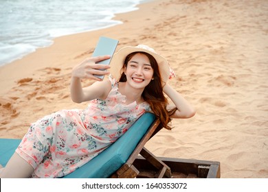 Relax Travel On The Beach Concept. Smile Asian Young Adult Woman Use Smart Phone Selfie For Social Network Connect. She Sit On The Beachchair Wear A Pink Dress And Sun Hat. 