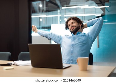 Relax At Work. Happy Bearded Man Sitting On Chair At Desk In Modern Office Using Laptop, Stretching His Back And Arms, Taking Break And Smiling, Enjoying His Job, Feeling Pleased And Satisfied