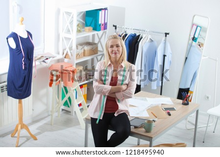 Relaxation after successful work. Unusual top view. Beautiful European woman looking at the camera, standing near the table in the workshop with clothes hanging in the background