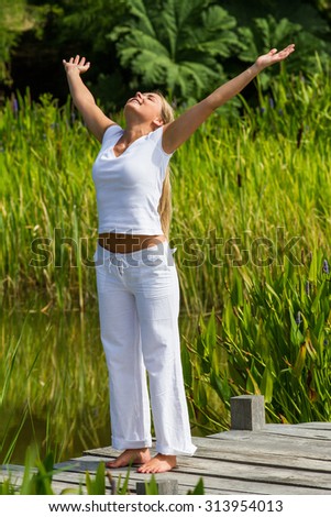 relaxation outside - relaxed young woman with bare feet outstretching her arms wide on a wooden path with green reed environment, summer daylight