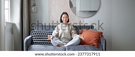 Relaxation and patience. Smiling young asian woman in cozy room, sitting on sofa and meditating, doing yoga mindfulness training.