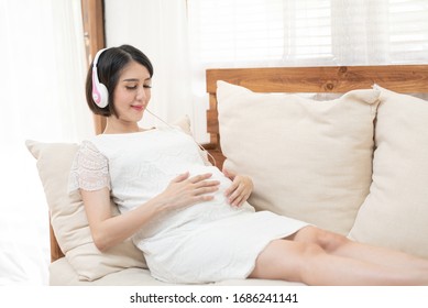 Relaxation in pregnancy. Calm pregnant young asian girl Listen to music Mozart effect good for fetus by using headphones attached to the stomach hold earphones near belly lying on sofa at home. - Powered by Shutterstock