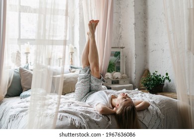 Relaxation, recreation concept. Dreamy and smiling young adult woman in pajamas raised her legs up lying on comfort bed in boho interior bedroom. Happy female spending early morning at home