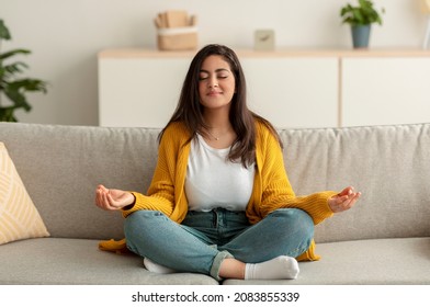 Relaxation, stress relief concept. Peaceful arab woman sitting in lotus position on couch in living room, meditating with closed eyes, copy space. Young lady enjoying weekend at home