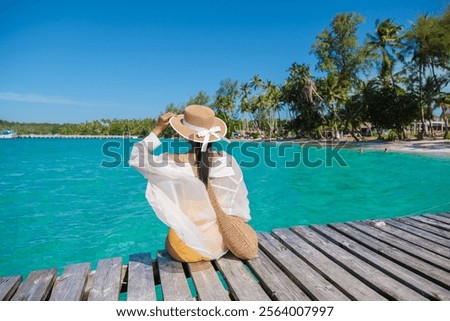 Relaxation unfolds as a woman enjoys the warm sun on her back while gazing at the turquoise waters of Koh Kood, surrounded by lush palms and tranquil scenery in this tropical paradise.