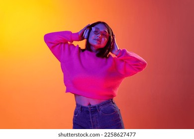 Relaxation  Young girl in cozy sweater listening to music in headphones isolated over gradient orange background in neon light  Concept emotions  facial expression  youth  inspiration  sales  ad
