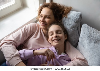 Relaxed affectionate family young adult single mom and cute teenage daughter resting hugging lying on bed together. Smiling mum embracing school kid girl bonding cuddling, lounge in bedroom, top view.