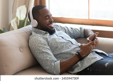 Relaxed african american man wearing wireless headphones listening music on smartphone sit on couch, happy young black guy holding phone using player app enjoy audio course or new songs at home