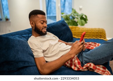 Relaxed african american smiling man sits on couch looks at smartphone at home in living room. Happy interested black guy laughing at funny video or joke, chatting with friends in social networks. - Shutterstock ID 2293334053