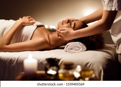 Relaxed African American Woman Enjoying Face Massage In Dark Spa Room