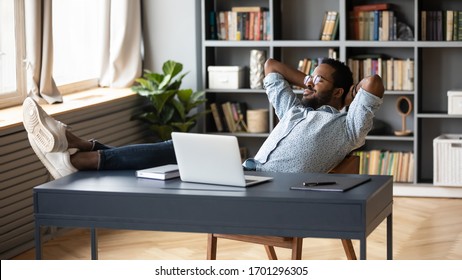 Relaxed african American young male sit at desk distracted from computer work take break daydreaming, calm biracial man lean in chair relax rest at office desk, breathe fresh air, stress free concept