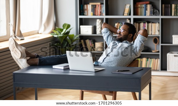 Relaxed african American young male sit at desk\
distracted from computer work take break daydreaming, calm biracial\
man lean in chair relax rest at office desk, breathe fresh air,\
stress free concept