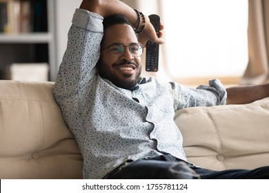 Relaxed African fellow resting on couch holding remote control watching tv shows television programs or favourite series movie, football fan enjoy match sport game, weekend lazy day at home concept