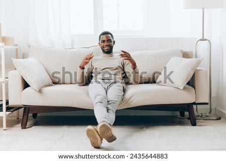 Relaxed AfricanAmerican man sitting on a comfortable sofa in a modern living room He's smiling, holding a pillow, and thinking while enjoying a relaxing weekend at home The background is minimalist,