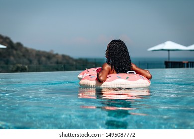 Relaxed African-American woman with inflatable donut in infinity pool, back view; blurred coastal landscape behind her.
