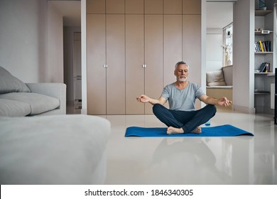 Relaxed aged man meditating with his eyes closed while sitting on a blue workout mat at home