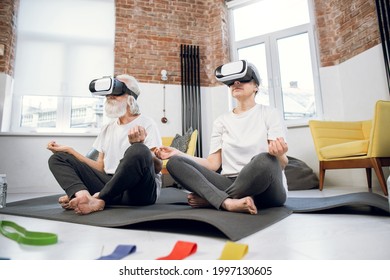 Relaxed Aged Man And Woman Wearing VR Glasses While Sitting In Lotus Position At Bright Living Room. Active Senior Couple Meditating At Home Using Innovative Headset.