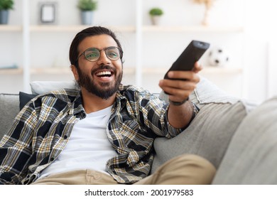 Relaxed Arab Guy Wathcing Movie On TV At Home, Sitting On Couch Alone, Having Happy Face Expression. Happy Young Man With TV Remote Watching Comedy And Laughing, Copy Space, Closeup