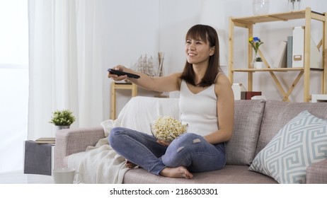 Relaxed Asian Chinese Woman Switching On Television And Laughing Loudly At Hilarious Talk Show While Eating Popcorn On Sofa