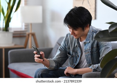 Relaxed Asian Man Using Mobile Phone While Sitting On A Couch At Home On A Daytime.