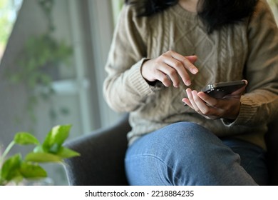 Relaxed Asian Woman In Comfy Sweater Using Her Smartphone, Searching Something On The Internet, Scrolling On Her Social Media While Sitting On Armchair In The Cafe. Cropped Image