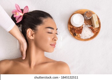 Relaxed Asian Woman With Flower In Hair Enjoying Neck Massage With Natural Ingredients Cosmetics At Spa, Lying On Table In Salon With Closed Eyes, Pampering Herself With Beauty Treatments, Top View