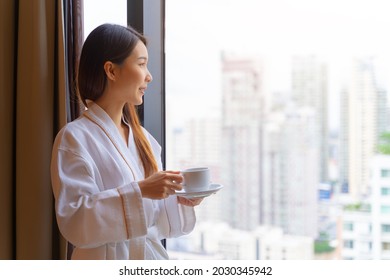 Relaxed Asian Woman Wearing Bath Robe Drinking Coffee In Hotel Room In Morning 