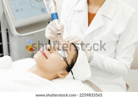 Relaxed Asian woman wearing protection glasses getting laser skin procedures in beauty salon. Clinic receiving face treatment. Beauty injection for Skincare. Close up of Beautician Hands.
