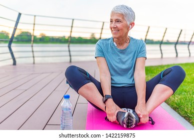 Relaxed Athletic Mature Woman Sitting On Fitness Mat Outdoors. Senior Woman Resting After Exercises. Woman On A Yoga Mat To Relax Outdoor. Senior Lady Prefers Healthy Lifestyle