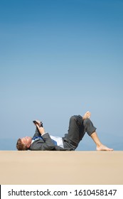 Relaxed Barefoot Businessman Lying On A Sunny Dock By The Sea Using His Smartphone