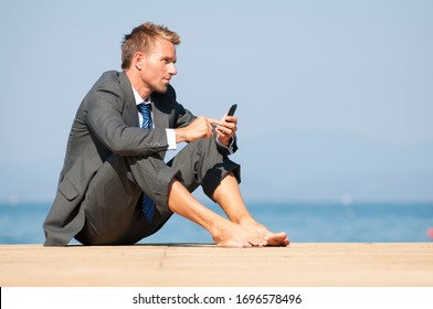 Relaxed Barefoot Businessman Sitting On A Sunny Dock By The Sea Using His Mobile Phone