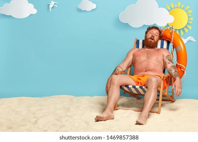 Relaxed Bearded Man Sleeps On His Deck Chair, Poses On Beach, Has Red Sunburned Skin, Sunbathes And Tans, Has Summer Vacation And Holiday Travel, Beautiful Sea View In Background. People And Rest