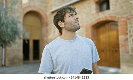 Relaxed, bearded young man, engrossed in his smartphone, gazes at the city sky in a green park - a portrait of a handsome adult immersed in texting, technology linking him to the world outside - Powered by Shutterstock