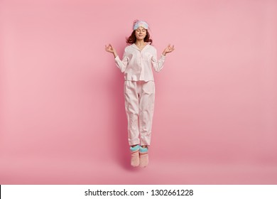 Relaxed beautiful woman meditates before having sleep, dressed in cotton pyjamas, warm chuni, eyemask, jumps against pink background, enjoys calm domestic atmosphere. Restful girl in nightsuit - Powered by Shutterstock