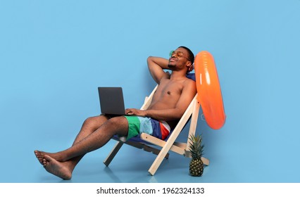 Relaxed black guy chilling in lounge chair with laptop computer, working online from tropical paradise, blue background. Cool young man on summer vacation watching movie or talking to friend on pc