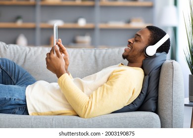 Relaxed Black Guy Laying On Couch, Watching Movies On Pad At Home, Copy Space. Happy African American Young Man Reclining On Sofa, Using Modern Digital Tablet And Wireless Headphones, Side View