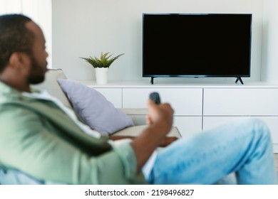 Relaxed Black Guy Watching TV Holding Remote Control And Switching Channels On Flatscreen Plasma Television Set With Blank Screen, Sitting On Sofa At Home, Focus On Tv