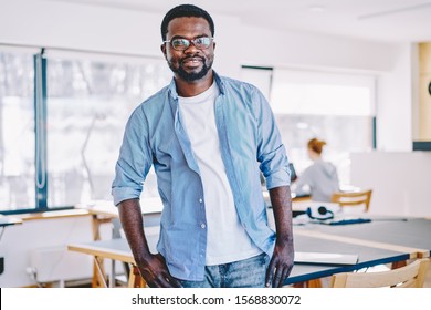 Relaxed black man in casual outfit smiling and looking at camera while leaning on table during break in spacious contemporary office