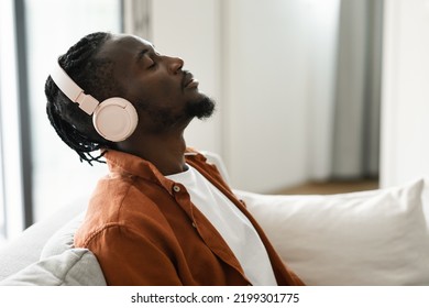 Relaxed Black Man In Headphones Sitting On Sofa With Closed Eyes, Listening To Music At Home, Side View, Copy Space. African American Male Enjoying New Audio Book In Headset