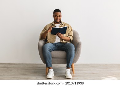 Relaxed Black Man Reading Book Sitting In Comfortable Armchair On Gray Background Indoors. African American Millennial Guy Reads Business Literature At Home. Weekend Leisure Concept