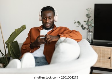 Relaxed Black Man Watching Movies On Smartphone, Wearing Headset And Using Cellphone, Sitting On Sofa, Copy Space. African American Guy Surfing Internet And Listening To Music At Home