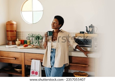 Relaxed black woman with afro hair standing in a kitchen, holding a cup of hot Brazilian coffee. She looks away thoughtfully, enjoying a hot drink in a cozy, homey setting.