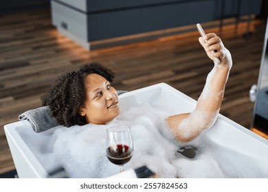 Relaxed Black woman lying in warm bubbly bath with glass of red wine in hand while video calling on phone in hotel bathroom, copy space - Powered by Shutterstock