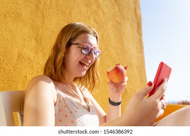 Relaxed Blond Teen Laughing While Eating A Red Apple, Lying In The Terrace Of The Resort And Making A Video Call In Her Smart Phone. Healthy Woman With Glasses Dieting, Biting Fruit In The Balcony