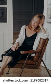 Relaxed Blond Woman In Black Evening Dress And White Shirt. Stylish Confident Mid Adult Lady Is Sitting In An Arm Chair. Happy European Woman Is Sitting Have Her Head Leaned To The Hand After Party.