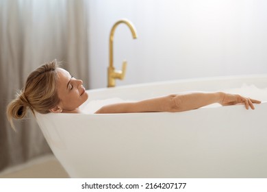 Relaxed Blonde Woman Enjoying Hot Foamy Aromatic Bath At Home, Laying In Tub Full Of Water With Closed Eyes, Side View, Closeup Photo, Copy Space. Home Spa, Body Care Concept