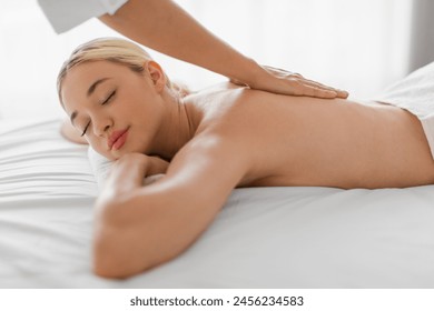 Relaxed blonde woman lies on a massage table at a spa, receiving a back massage from a massage therapist. The therapist is using firm pressure and various techniques - Powered by Shutterstock