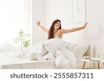 Relaxed brunette girl with smile on her face stretching after waking up in cozy bed. Happy young woman sitting under white blanket in bright sunlit bedroom, savoring peaceful weekend morning at home.