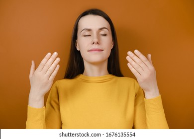 Relaxed Brunette Woman Enjoying Good Smell Or Pleasant Fragrance, Serene Mindful Young Female Taking Deep Breath Feel No Stress Free Inhaling Fresh Air Relaxing Isolated On Orange Studio Background