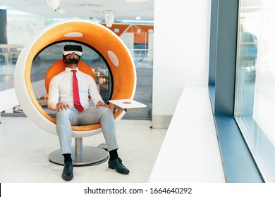 Un relajado hombre de negocios disfruta de un vídeo virtual en el moderno salón de oficinas. Hombre con ropa de oficina y lentes de realidad virtual sentado en una silla interactiva y mirando hacia arriba. Relajación del concepto de oficina